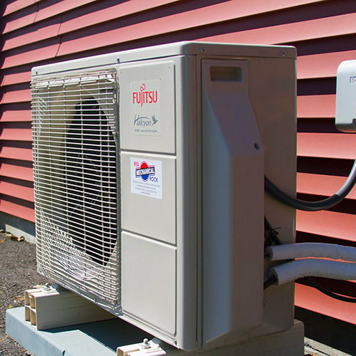 Heat pump condenser with red siding background.