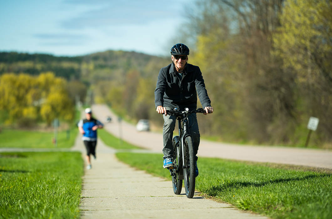 Man riding electric bike towards camera on spring day with runner and green grass in the background.