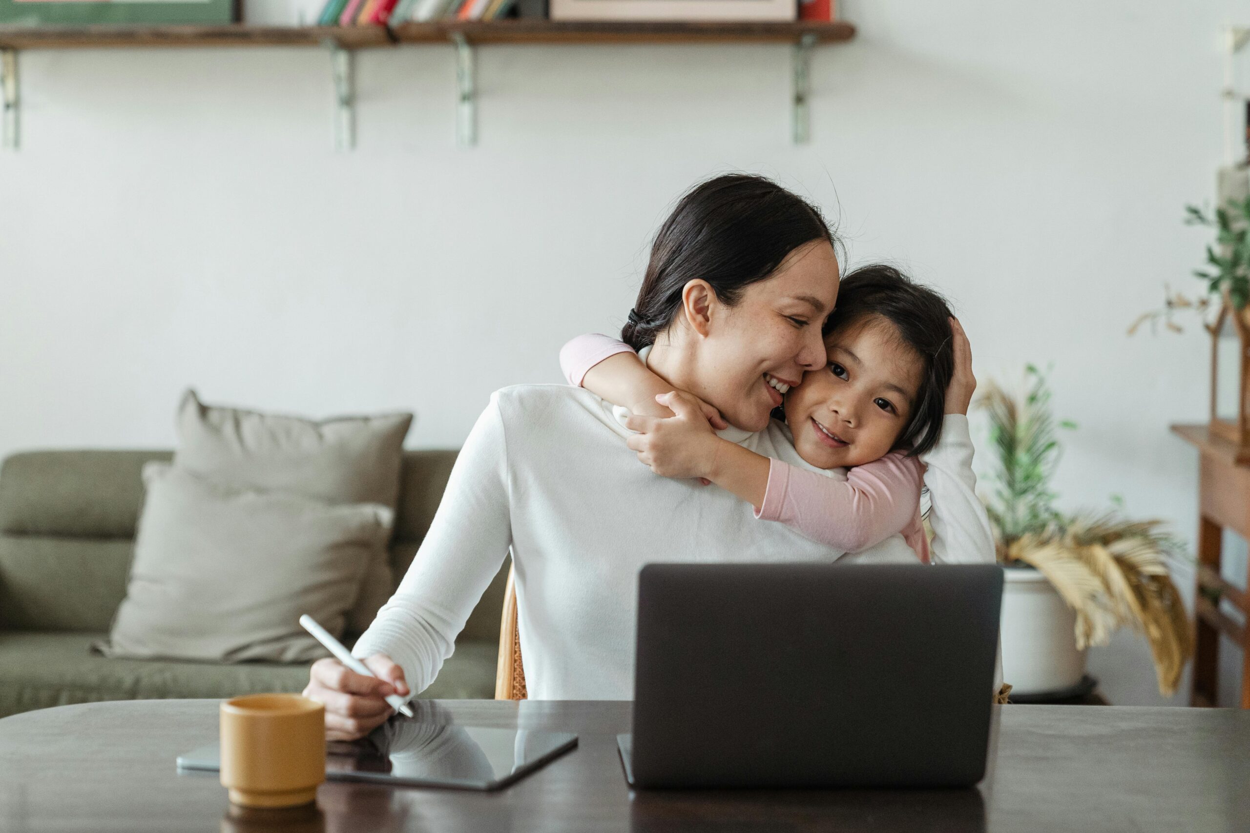 Mom in white shirt and dark hair with laptop and tablet in front of her, half embracing young child who is hugging her neck