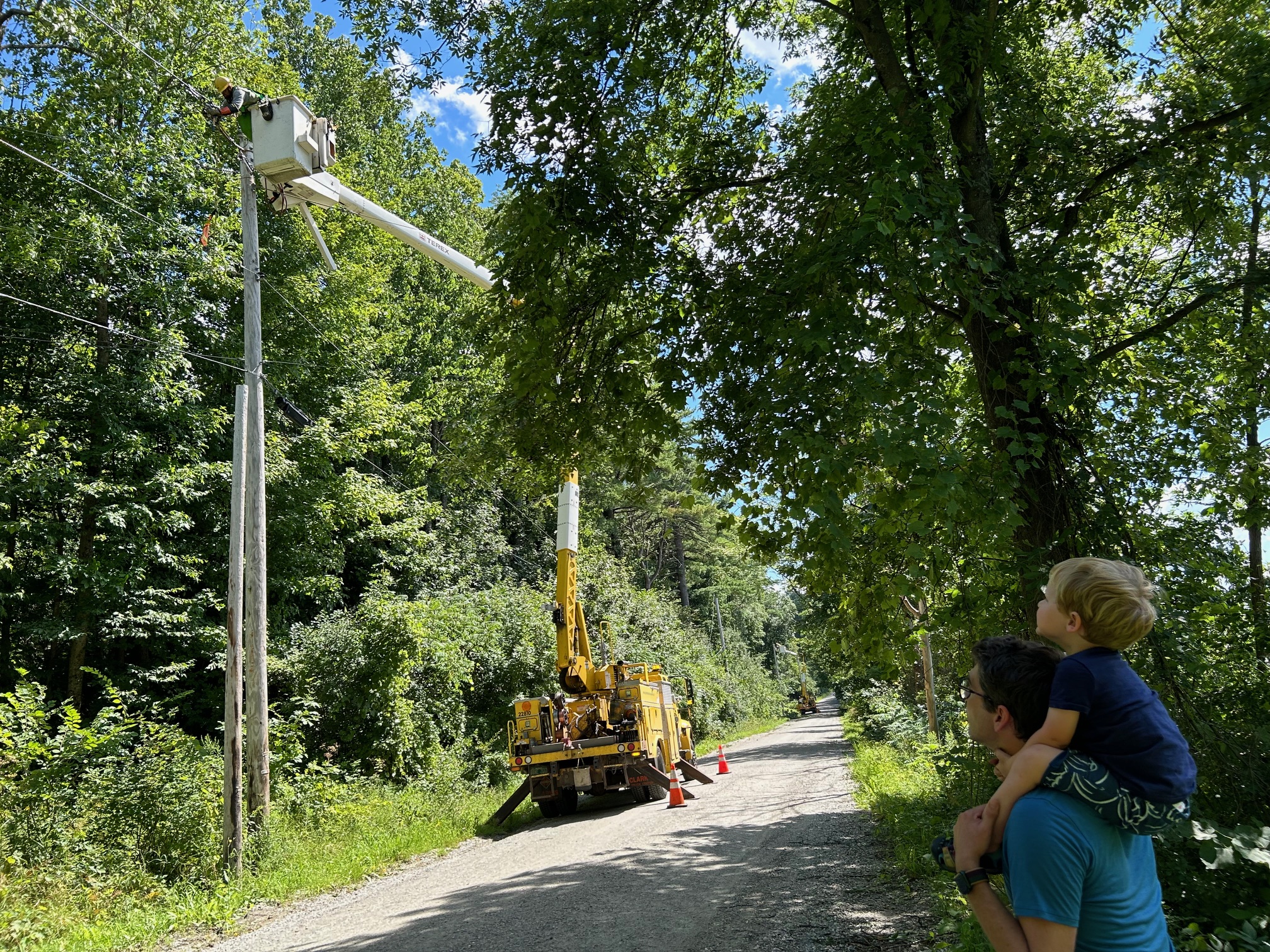 Adult with child on shoulders, watching GMP workers work on power lines on a dirt road with many trees around