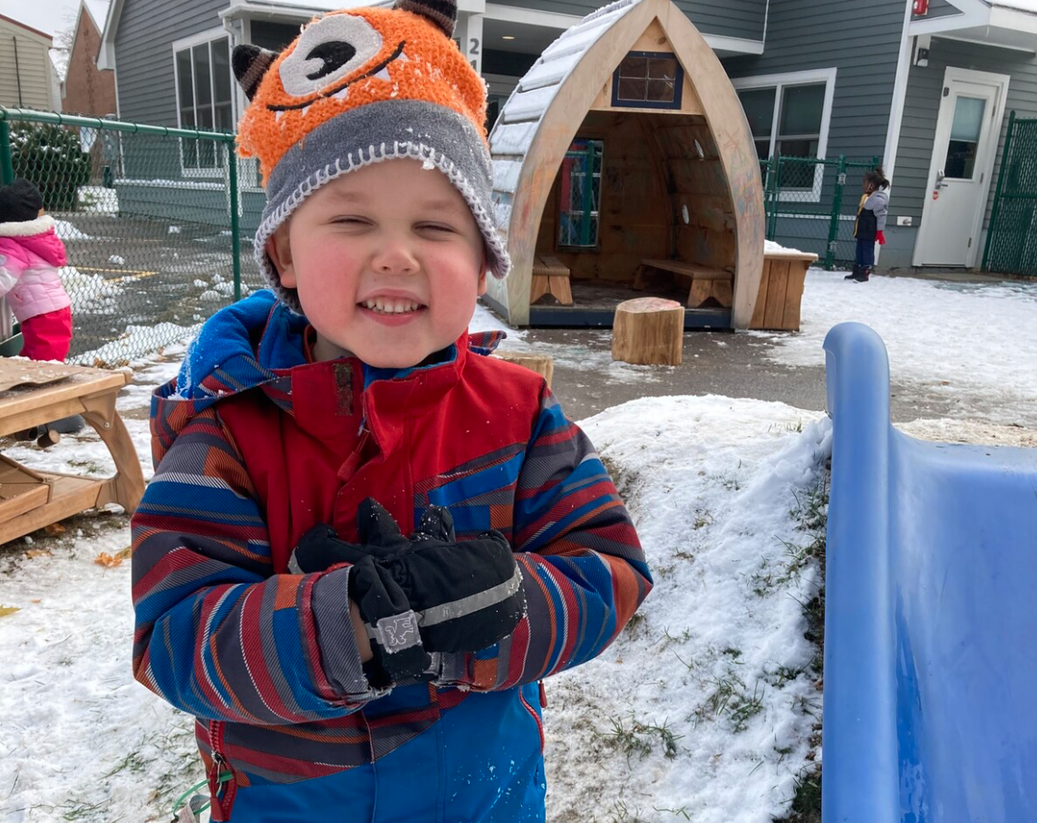 Young child, wearing a multi-color jacket, mittens and a monster hat, standing in the snow