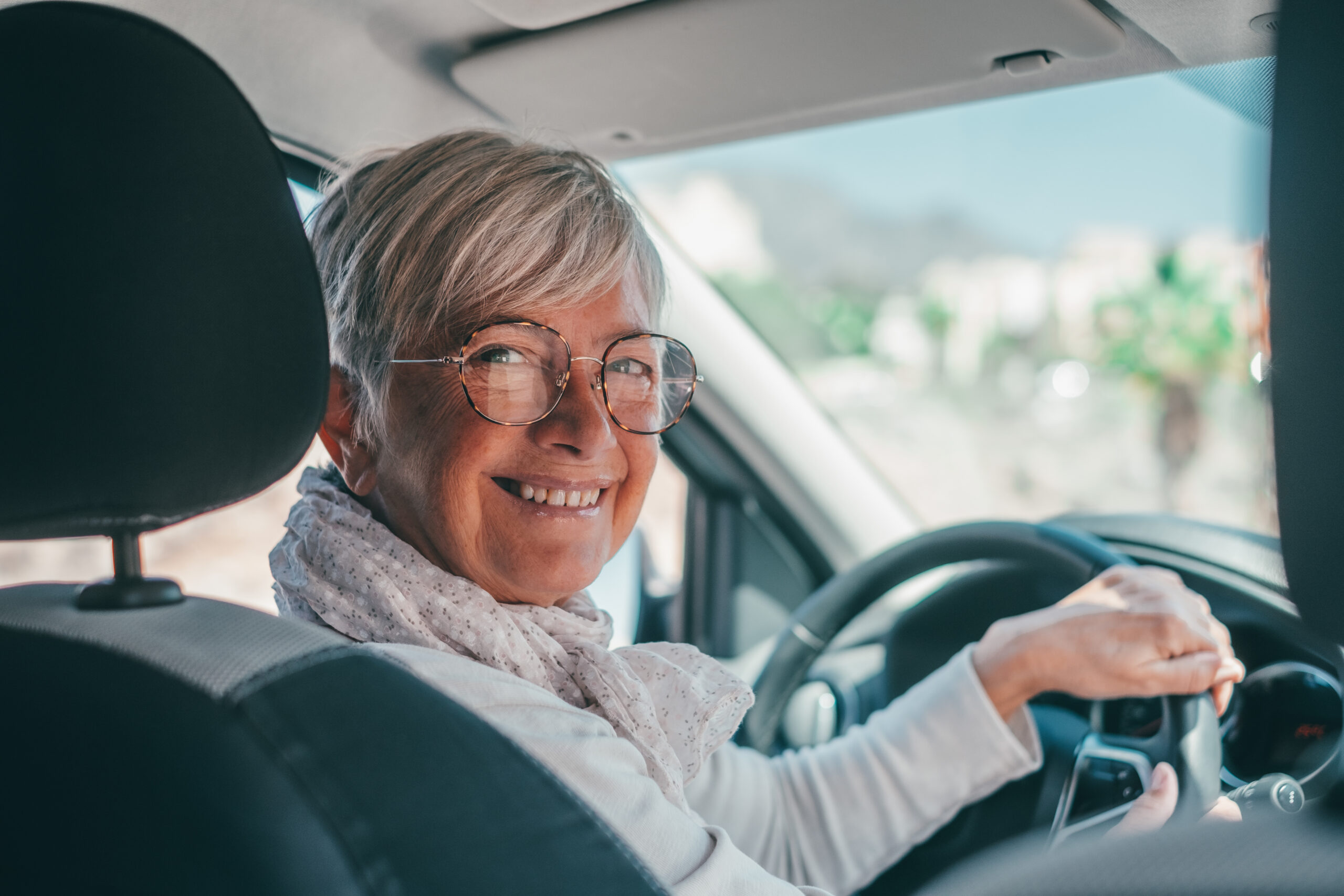 A woman with short, white hair and glasses, sitting behind the wheel of a car and smiling back at the camera